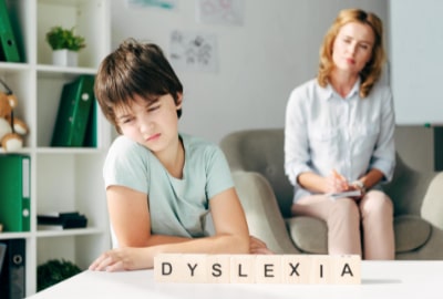 a dyslexic boy kneeling at the table looking dejecting with his mother sitting in the background on the sofa
