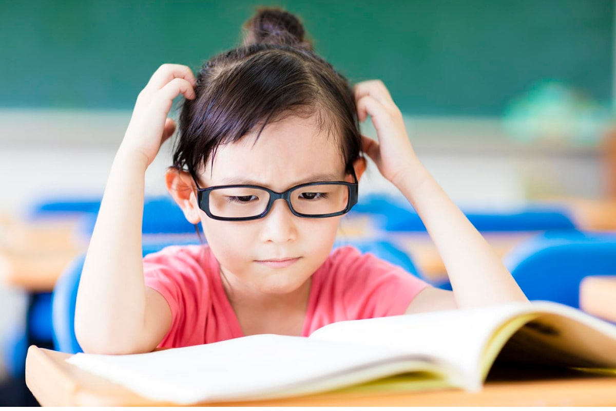 Featured image for the post on why some kids struggle with reading. The image shows a young girl with glasses, sitting at her desk and feeling frustrated because she can't read well